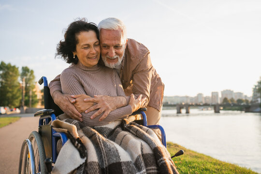 Senior woman in wheelchair walking with caregiver old man on road in park. Elderly family couple man supporting embracing woman in chair for people with disability outdoor. Rehabilitation