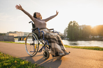 Happy senior woman in wheelchair raised hands enjoying freedom walking on road in city park outdoor. Old happy woman in chair for people with disability has a hope. Disability disease rehabilitation