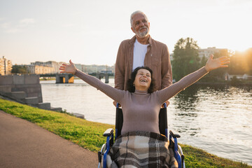 Senior woman in wheelchair walking with caregiver old man on road. Family couple man husband supporting taking care of paralyzed woman wife in chair for people with disability outdoor. Rehabilitation
