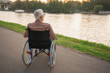 Sad senior man sitting in wheelchair walking on road in city park outdoor, back view. Old paralyzed man in chair for people with disability feel depressed. Lonely elderly sick man sits on wheelchair