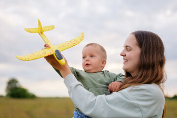 Smiling mother holds baby boy and toy airplane, enjoying playful outdoor moments in a scenic field under a cloudy sky.
