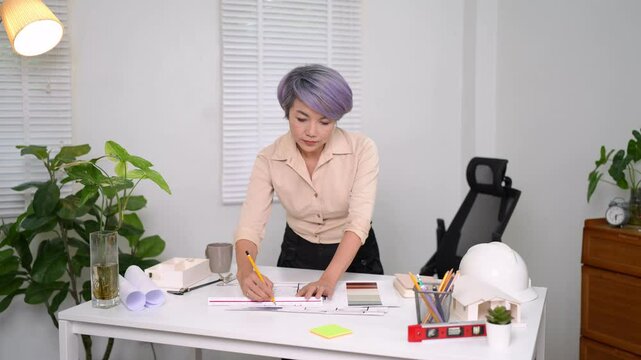 Senior Asian female architect in beige shirt drawing with pen while standing at bright office desk
