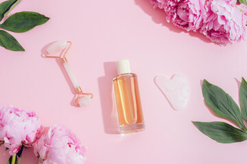Cosmetic bottle, face roller, gua sha stone and pink peonies on pastel pink background, skincare concept. Top view, flat lay