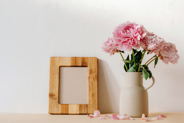 Pink peonies in ceramic jug and blank wooden picture frame against white wall. Mockup