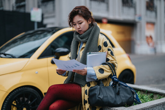 A stylish young adult sits by a yellow car holding pages and notebooks, indicating thoughtful analysis, business preparation, or academic study while maintaining a modern urban vibe.
