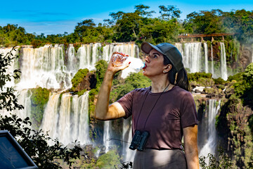 A female tourist drinks water while exploring Iguazu Falls on a sunny day. Wearing a cap and...