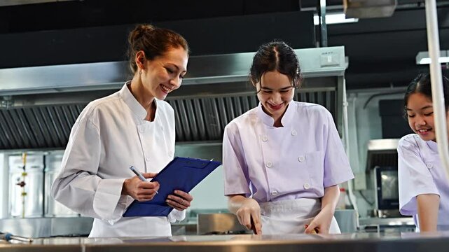 Female chef teacher teaches two students to cook in class. A cooking test is given to the teenage girls to cook spaghetti. The teacher holds a file to give scores in the cooking class.