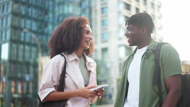 Woman and man having conversation outdoors, woman holding smartphone with expressive face, man gesturing enthusiastically. Cityscape backdrop. Mixed-races couple talking at street and smiling.