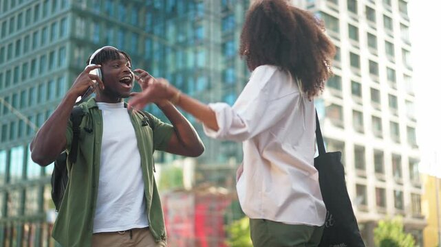 Man wearing headphones and holding a smartphone, gesturing in surprise during an unexpected encounter with a woman. Couple of friends meeting randomly at street. Random meet in city.