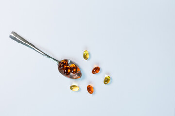 A close-up, top-view image of golden fish oil capsules on a silver spoon, isolated on a clean white background. This flat lay composition represents the concept of daily nutritional supplements.