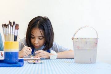 Young Asian girl focuses on painting a clay object, with her hand and brush with selective focus shows creativity and calm concentration for kid learning activity  via art and development in child.