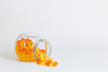Glass jar with yellow vitamin capsules spilled on white surface. Clean layout with copy space...