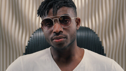 Portrait African American guy man ethnic male putting on sunglasses glasses wearing looking at camera sitting in futuristic white studio futurism innovation stylish fashion posing indoors sit in chair