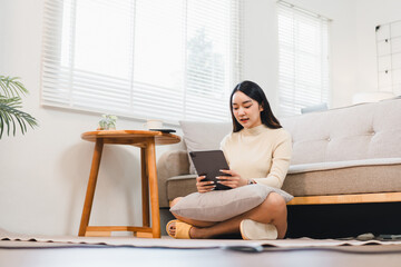 Young Asian woman sitting on floor in cozy living room, using digital tablet with relaxed and focused expression, natural light from window, modern home interior