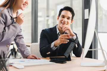 Businessman and businesswoman discussing project at modern office desk, smiling and collaborating with digital tablet and computer, natural daylight creates positive atmosphere