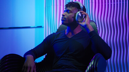 African American man guy male sitting in chair listening music in headphones using wireless earphones listen audio enjoy song sound enjoy modern cyber technology neon futurism room futuristic violet