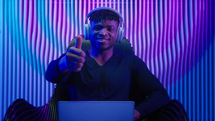 African American man guy male coder gamer programmer in chair wear headphones using laptop online work crypto software play internet game modern technology neon futurism showing thumb up like gesture