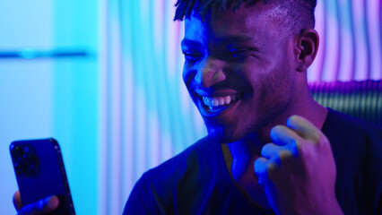 African American man guy male indoors futuristic innovation cyberspace neon lights winning with mobile phone bet casino online crypto win victory champion celebrate yelling happy video game addict