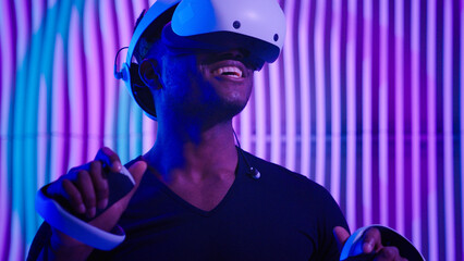 Excited African American man guy male gamer gaming vr play virtual reality glasses helmet internet game modern cyber technology neon futurism indoors futuristic metaverse playing software AR designer