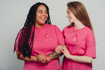 Two smiling women supporting breast cancer awareness, holding hands and wearing pink ribbons