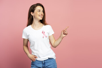 Young woman pointing aside, wearing pink ribbon for breast cancer awareness, smiling