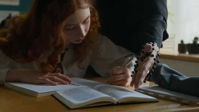 Slowmo shot of concentrated teen Caucasian girl in uniform studying at desk in religious school with unrecognizable nun teacher holding rosary in hands helping her with assignment