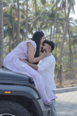 A cheerful Indian couple shares a romantic moment on a palm-lined road, standing in front of a black SUV. The woman is wearing a flowing lavender gown, and the man embraces her from behind with a joyf