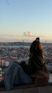 Young woman sitting on a wall, admiring the panoramic cityscape of lisbon, portugal, with the iconic 25 de abril bridge in the background, during a beautiful sunset. Vertical video