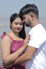 A cheerful Indian couple enjoys a sunny day at the beach, holding hands while walking along the shoreline. The woman is dressed in a casual maroon dress, and the man wears a white t-shirt with grey sh