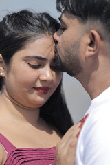 A cheerful Indian couple enjoys a sunny day at the beach, holding hands while walking along the shoreline. The woman is dressed in a casual maroon dress, and the man wears a white t-shirt with grey sh