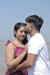 Obraz premium A cheerful Indian couple enjoys a sunny day at the beach, holding hands while walking along the shoreline. The woman is dressed in a casual maroon dress, and the man wears a white t-shirt with grey sh