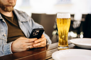 Man drinking beer from glass in cafe while using smartphone.