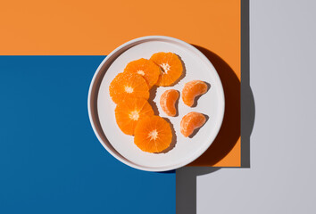 Rodajas y gajos de mandarina en un plato blanco sobre fondo naranja y azul. Vista superior