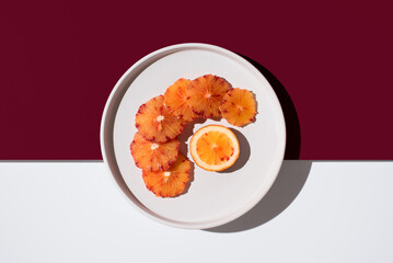 Rodajas de naranja sanguina sobre un plato blanco con fondo blanco y granate. Vista superior