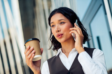 Beautiful businesswoman outdoors, calling on a phone while holding coffee, exemplifies...