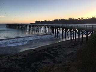 Obraz premium A picturesque wooden pier stretches into the Pacific Ocean at San Simeon, California, bathed in the warm glow of a sunset, inviting tranquility.