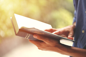 A close-up of a man reading the Holy Bible outdoors in natural sunlight, symbolizing faith,...