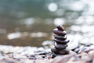 Obraz premium Stack of smooth black pebbles balanced on a rocky riverbank with blurred water background. Represents balance, meditation, harmony, nature, and mindfulness in a peaceful outdoor setting.