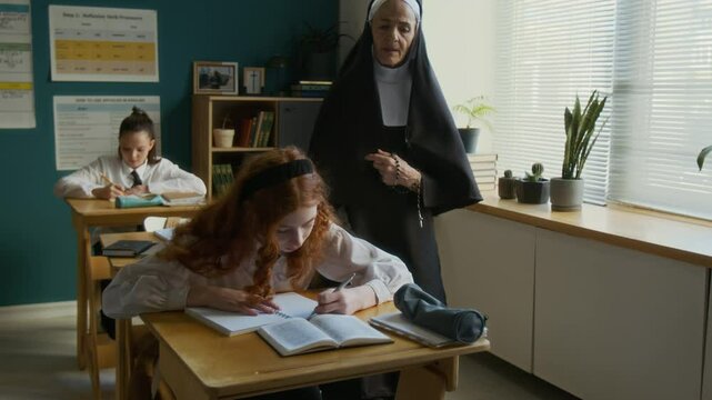 Mature Caucasian nun teacher in veil checking multiethnic student girls assignment during catechism class in religious school