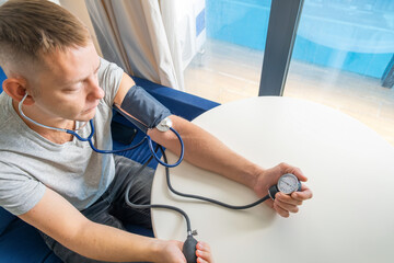 Man using manual sphygmomanometer and stethoscope to check his own blood pressure while sitting at home. Managing hypertension, heart health and general wellness. Regular self health monitoring