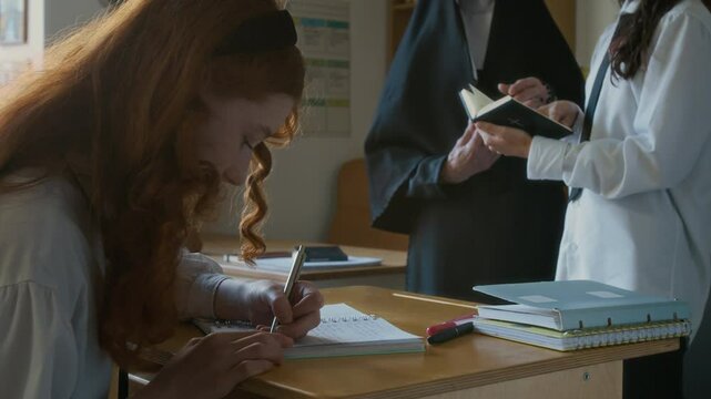 Focused teen Caucasian student girl writing in notebook at school desk while her classmate reading Bible with nun teacher standing at blackboard in front of class