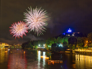 Fireworks over the Po River in Turin, Italy, during the San Giovanni celebration, with illuminated boats, Monte dei Cappuccini, and the Church of Santa Maria al Monte lit in blue.
