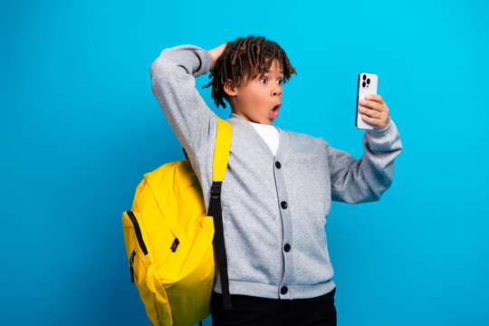 Surprised schoolboy with a bright yellow backpack looking at his smartphone against a vibrant blue background
