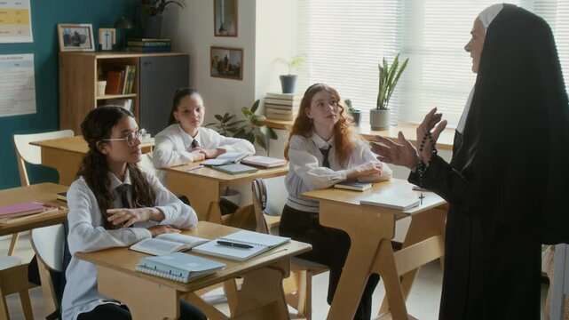 Motivated teen Caucasian student girl raising hand during class studying in religious school with senior nun in veil leading lesson