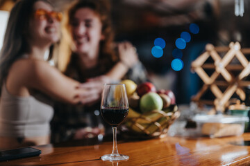 A glass of red wine on a wooden countertop with blurred individuals enjoying a lively gathering, showcasing a cozy social setting.