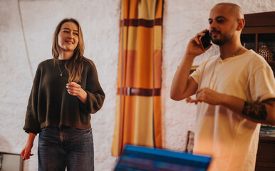 Two individuals engage in a casual, indoor social setting, one talking on a phone while another smiles, creating a relaxed scene showcasing communication and interactions.