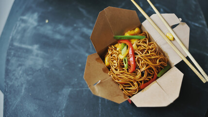 A high-quality food photograph of Chinese lo mein noodles served in a brown paper takeout container with wooden chopsticks