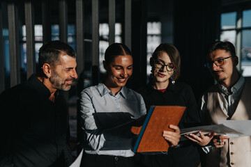 Naklejka na ściany i meble Group of diverse business people happily discussing ideas while gathered around a tablet in a modern office environment. They exhibit teamwork and creativity while engaging in a collaborative project.