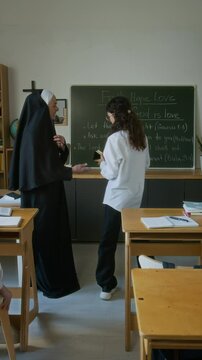Vertical of motivated teen Caucasian student girl raising hand during class in religious school and answering at blackboard in front of senior nun teacher and classmates