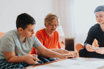 A grandmother enjoying a card game with her grandchildren at a cozy living room table. The family...
